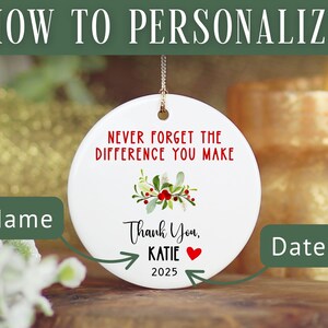 May include: White ceramic ornament with the text "NEVER FORGET THE DIFFERENCE YOU MAKE" in red. It includes a floral design and the words "Thank You, KATIE 2025". The ornament is designed to be personalized.