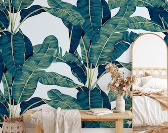 Op de afbeelding: Een slaapkamer met een behang met tropische bladeren in tinten groen en blauw. Een houten bank, een geweven vloerkleed en een decoratieve mand staan voor de muur. Een spiegel en een bed zijn ook zichtbaar.