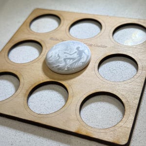 Könnte beinhalten: Ein Holz-Display-Brett mit neun runden Ausschnitten zum Aufbewahren von Knöpfen oder Abzeichen. Das Brett hat den Text "Rule Zero Tag Guide" aufgedruckt. Ein weißer Knopf mit einem schwarz-weißen grafischen Design ist in einem der Ausschnitte ausgestellt.