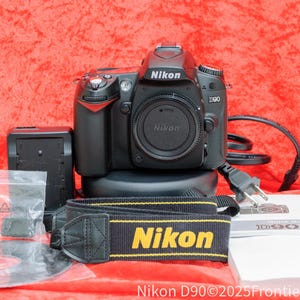 May include: A black Nikon D90 digital SLR camera with a lens attached. The camera is sitting on a black base with a black strap with yellow lettering that says "Nikon".