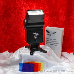 May include: A black Vivitar Auto Thyristor 2800 electronic flash with a white diffuser. The flash is on a black stand, with a booklet and coloured filters. The text "Vivitar 2800" is visible.