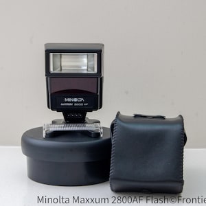 May include: A black Minolta Maxxum 2800AF flash unit with a black carrying case. The flash unit has a built-in diffuser and a swivel head.