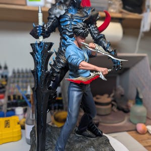 May include: A detailed collectible figurine featuring a young person in a blue hoodie and dark pants holding two daggers, standing in front of a large, dark, armored knight figure with a long black sword and a flowing red plume on a rocky base.