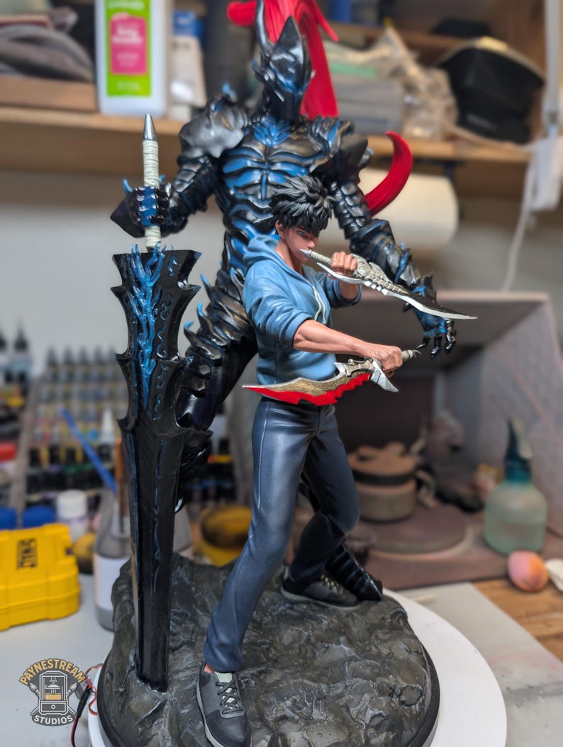 May include: A detailed collectible figurine featuring a young person in a blue hoodie and dark pants holding two daggers, standing in front of a large, dark, armored knight figure with a long black sword and a flowing red plume on a rocky base.