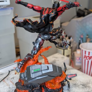 K&ouml;nnte beinhalten: Eine detaillierte Actionfigur eines geh&ouml;rnten Charakters mit roter und schwarzer Haut, der einen schwarzen Umhang tr&auml;gt und zwei rote Lichtschwerter schwingt. Die Figur ist in einem dynamischen Sprung &uuml;ber einem Sockel aus Lava dargestellt.