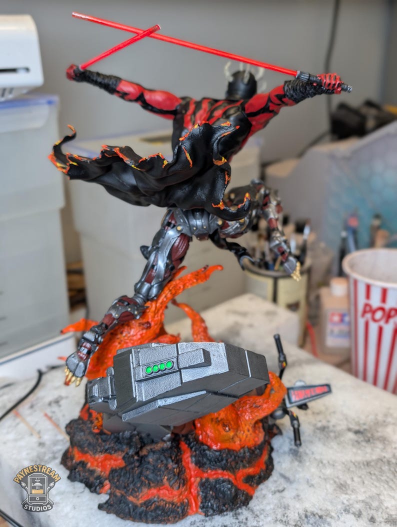 K&ouml;nnte beinhalten: Eine detaillierte Actionfigur eines geh&ouml;rnten Charakters mit roter und schwarzer Haut, der einen schwarzen Umhang tr&auml;gt und zwei rote Lichtschwerter schwingt. Die Figur ist in einem dynamischen Sprung &uuml;ber einem Sockel aus Lava dargestellt.