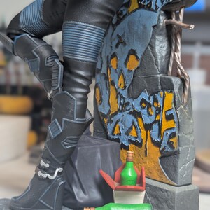 May include: A collectible figurine featuring a character in black pants with blue ribbed details, leaning against a stone wall with yellow and blue graffiti. Two green bottles and a red and white takeout container sit at the base of the structure.