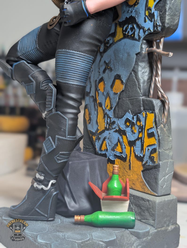 May include: A collectible figurine featuring a character in black pants with blue ribbed details, leaning against a stone wall with yellow and blue graffiti. Two green bottles and a red and white takeout container sit at the base of the structure.