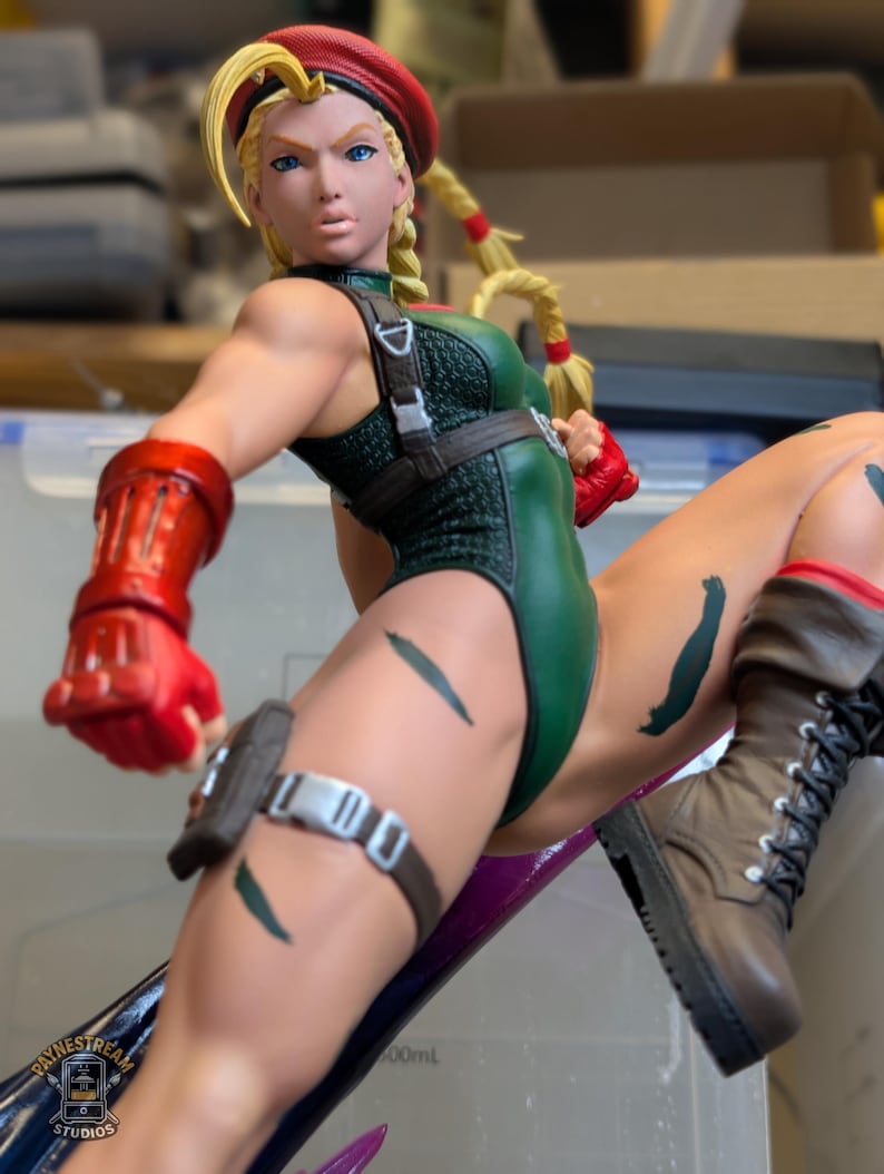 May include: A collectible figurine of a blonde character wearing a red beret, a green leotard with a textured pattern, red gauntlets, and brown combat boots. The character is in a dynamic pose with a brown utility belt and a logo reading Paynestream Studios.