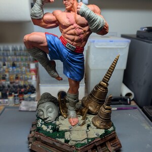 May include: A muscular, bald fighter figurine with a scarred chest, wearing blue shorts with a red waistband labeled TIGER and white hand wraps. The figure stands on a detailed base featuring a stone Buddha head and golden temple bells.