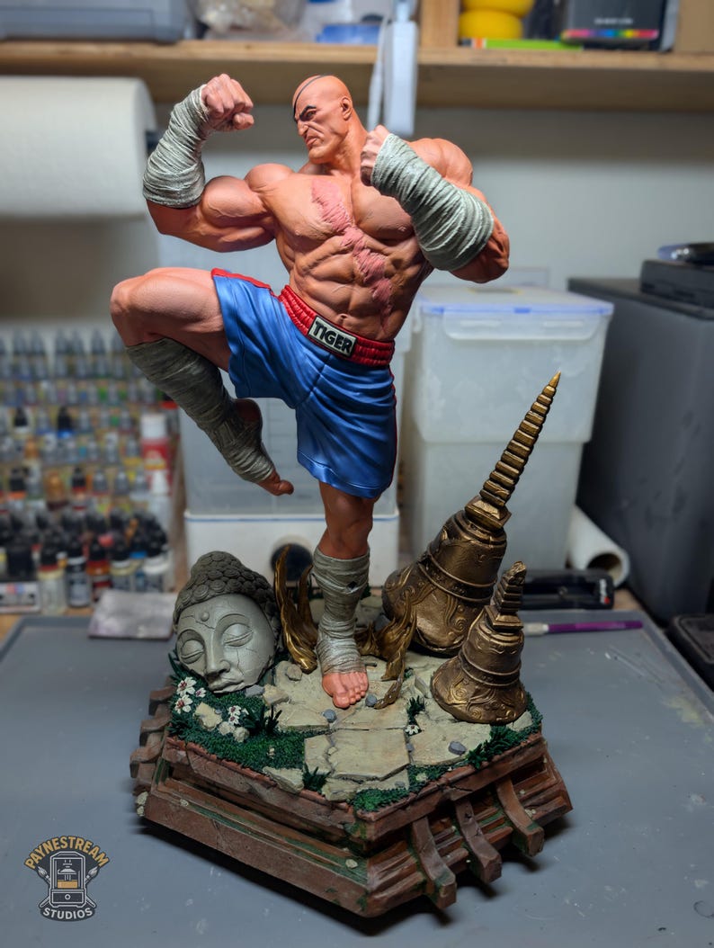 May include: A muscular, bald fighter figurine with a scarred chest, wearing blue shorts with a red waistband labeled TIGER and white hand wraps. The figure stands on a detailed base featuring a stone Buddha head and golden temple bells.
