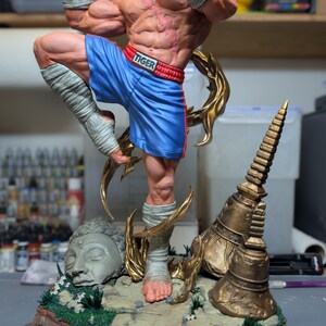 May include: A muscular male figurine in a dynamic fighting pose, wearing blue shorts with the word TIGER on the waistband and white hand wraps. The base features a stone Buddha head, golden temple bells, and green foliage on a rocky ground.