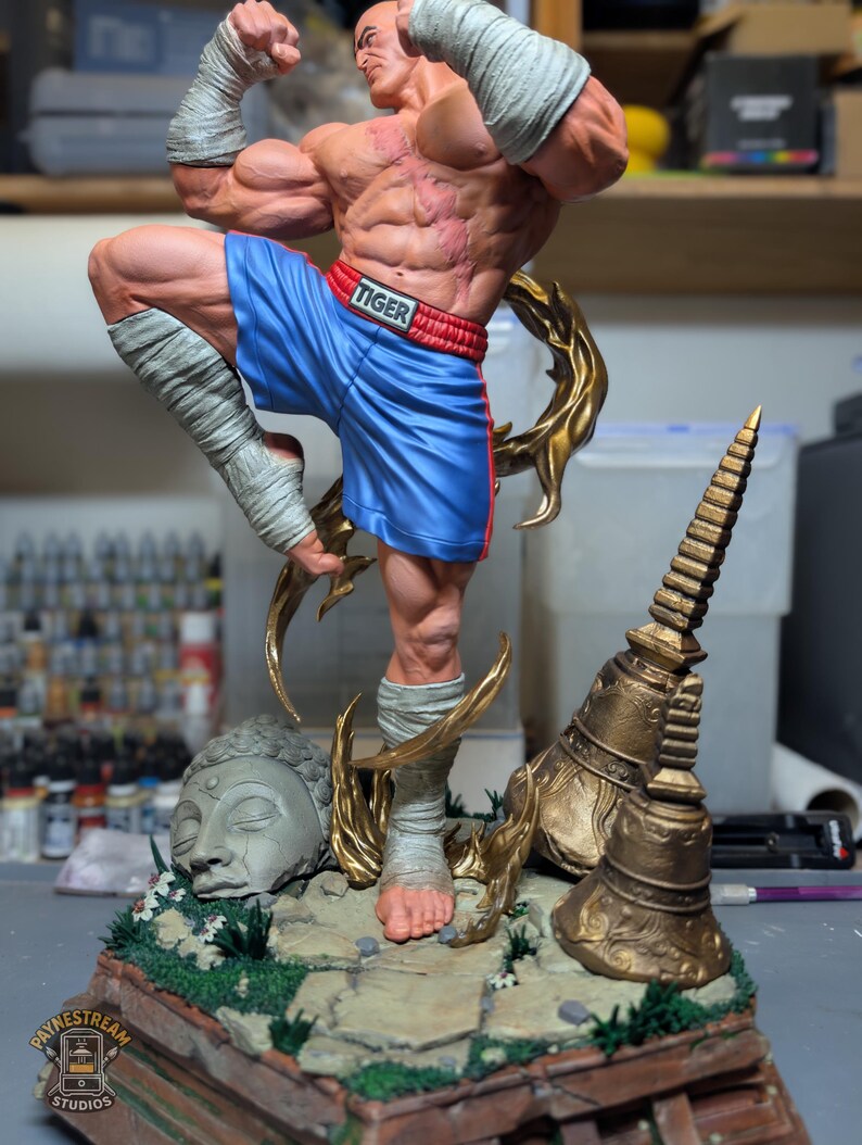May include: A muscular male figurine in a dynamic fighting pose, wearing blue shorts with the word TIGER on the waistband and white hand wraps. The base features a stone Buddha head, golden temple bells, and green foliage on a rocky ground.