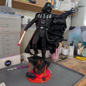 May include: A Darth Vader figurine standing on a rocky base with orange and red lava details. The character wears a black suit, cape, and helmet, with one hand outstretched and the other holding a glowing red lightsaber blade extending from the hilt.