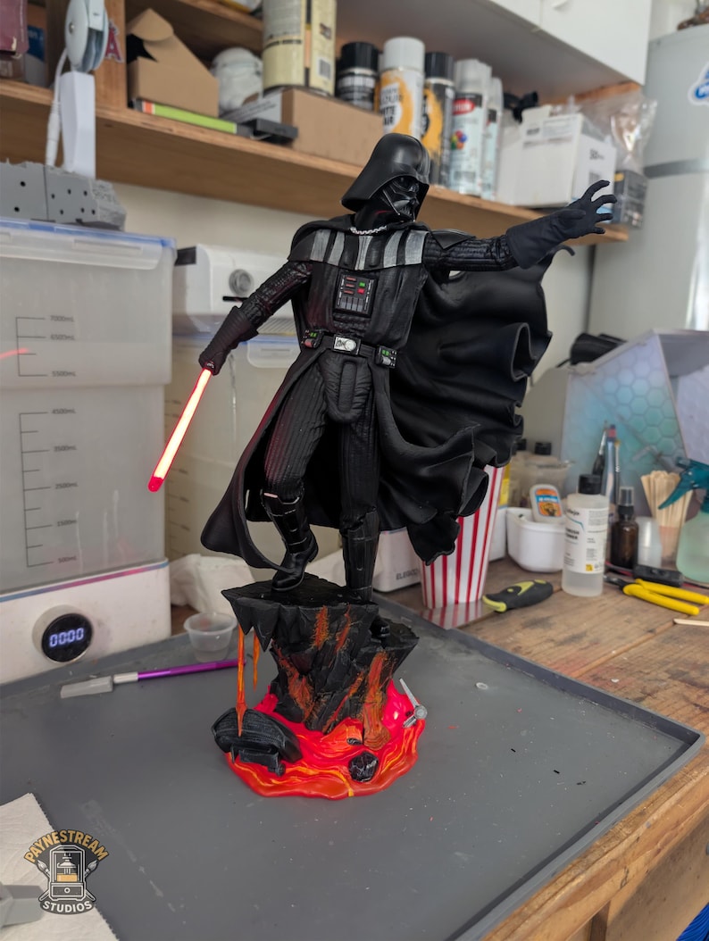 May include: A Darth Vader figurine standing on a rocky base with orange and red lava details. The character wears a black suit, cape, and helmet, with one hand outstretched and the other holding a glowing red lightsaber blade extending from the hilt.