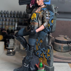 May include: A detailed figurine of a character with bright pink hair, wearing a brown jacket over a gray graphic crop top, black pants, and boots. The character holds a black rifle and leans against a weathered stone wall with graffiti.