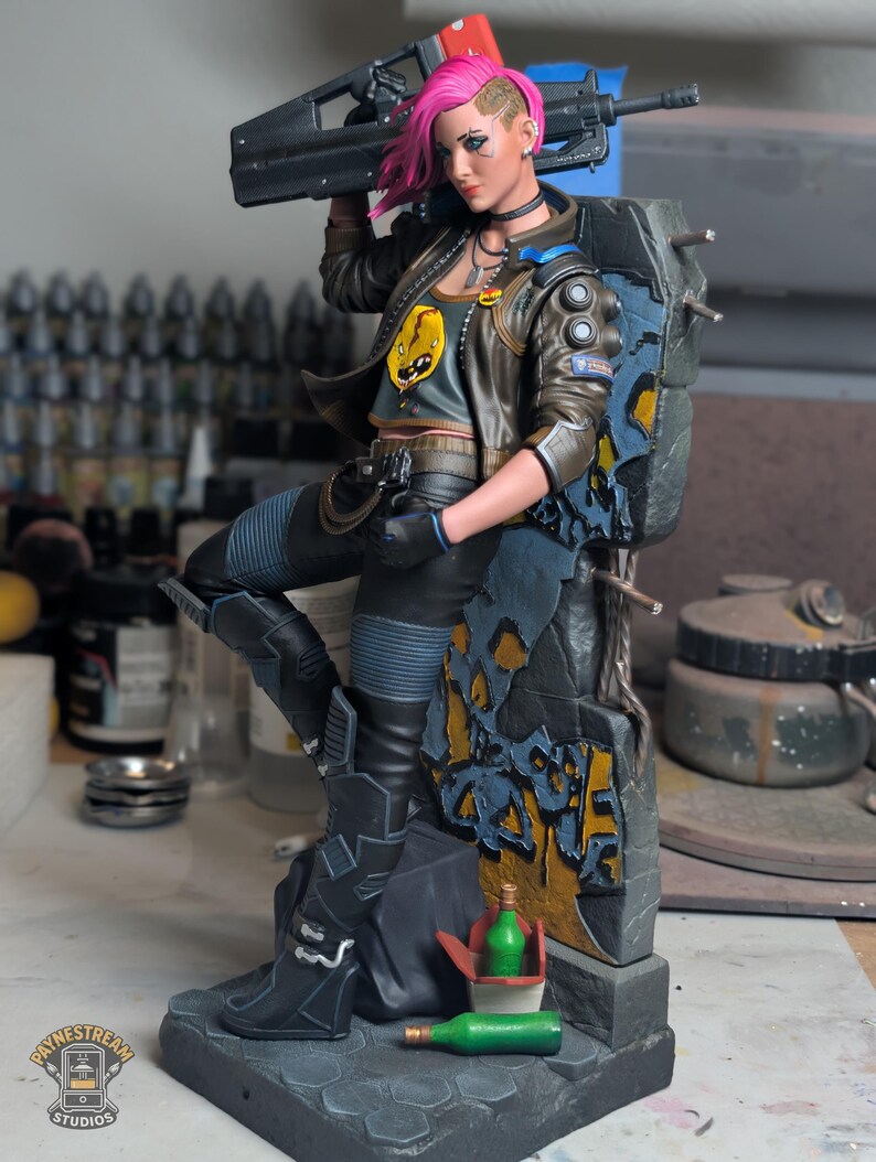 May include: A detailed figurine of a character with bright pink hair, wearing a brown jacket over a gray graphic crop top, black pants, and boots. The character holds a black rifle and leans against a weathered stone wall with graffiti.