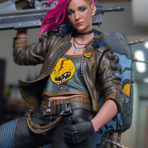 May include: A collectible action figure featuring a character with bright pink hair, wearing a brown leather jacket over a gray crop top with a yellow graphic, black pants with a gold chain, and black gloves, holding a large black rifle.