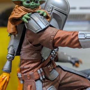 K&ouml;nnte beinhalten: Detaillierte Figur von The Mandalorian mit Grogu auf seiner Schulter. Der Mandalorianer tr&auml;gt eine braune und silberne R&uuml;stung, Grogu einen braunen Umhang. Die Figur hat eine dynamische Pose, mit einem sichtbaren Flammeneffekt.