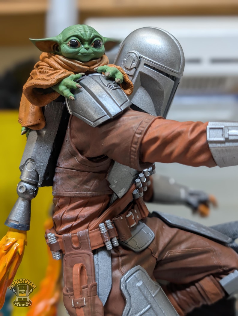 K&ouml;nnte beinhalten: Detaillierte Figur von The Mandalorian mit Grogu auf seiner Schulter. Der Mandalorianer tr&auml;gt eine braune und silberne R&uuml;stung, Grogu einen braunen Umhang. Die Figur hat eine dynamische Pose, mit einem sichtbaren Flammeneffekt.
