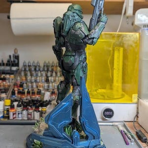 May include: A green armored soldier figurine standing on a blue geometric base, holding a long rifle pointed upward. The figure features detailed mechanical plating, a helmet, and a dark undersuit, presented in a standing pose on a textured surface.