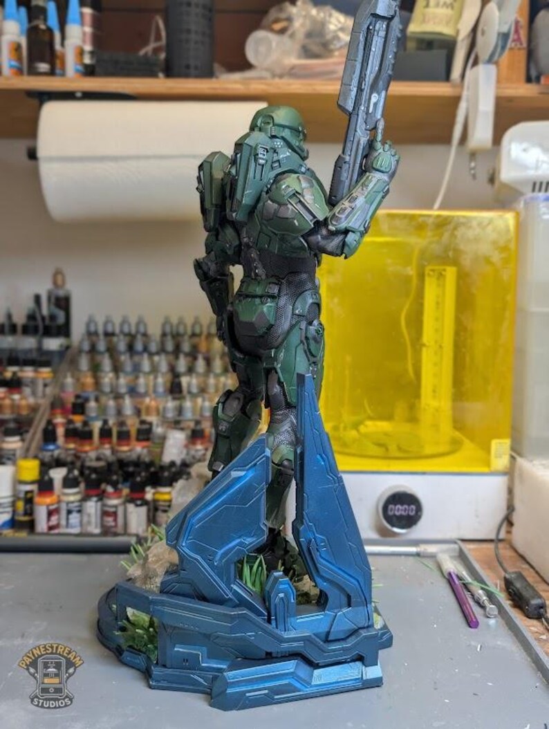 May include: A green armored soldier figurine standing on a blue geometric base, holding a long rifle pointed upward. The figure features detailed mechanical plating, a helmet, and a dark undersuit, presented in a standing pose on a textured surface.