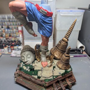 May include: A painted resin figurine of a martial artist in a dynamic pose, wearing blue boxing shorts with the word TIGER on the waistband. The figure stands on a detailed base featuring a stone Buddha head, golden temple bells, and greenery.