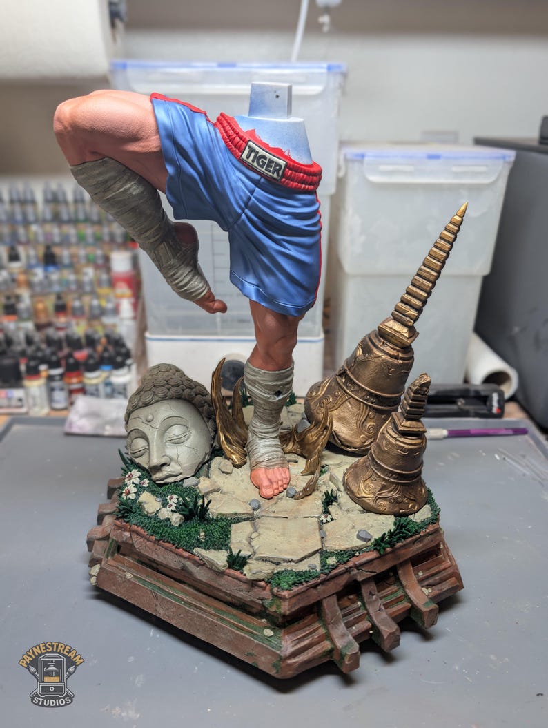 May include: A painted resin figurine of a martial artist in a dynamic pose, wearing blue boxing shorts with the word TIGER on the waistband. The figure stands on a detailed base featuring a stone Buddha head, golden temple bells, and greenery.