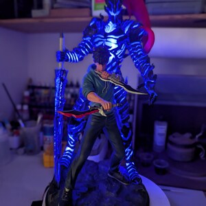 May include: A collectible figurine featuring a character in a blue hoodie and dark pants holding two daggers, standing in front of a glowing blue spectral warrior figure with a red ponytail and a large sword, all mounted on a dark rocky base.
