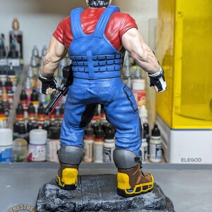 May include: A detailed figurine of a character wearing a red cap, red shirt, and blue overalls. The figure has a tactical vest, black gloves, and brown boots with yellow accents. It stands on a gray, textured base. The figure is holding a black weapon.