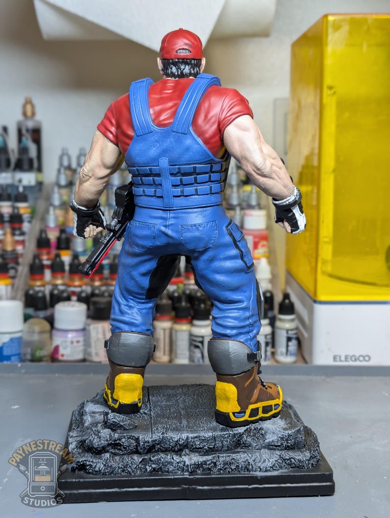 May include: A detailed figurine of a character wearing a red cap, red shirt, and blue overalls. The figure has a tactical vest, black gloves, and brown boots with yellow accents. It stands on a gray, textured base. The figure is holding a black weapon.