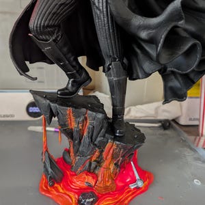 May include: A detailed figurine of a character wearing black ribbed trousers, a flowing black cape, and black boots, standing on a textured rock base with orange and red lava accents, featuring a glowing red lightsaber blade extending from the side.