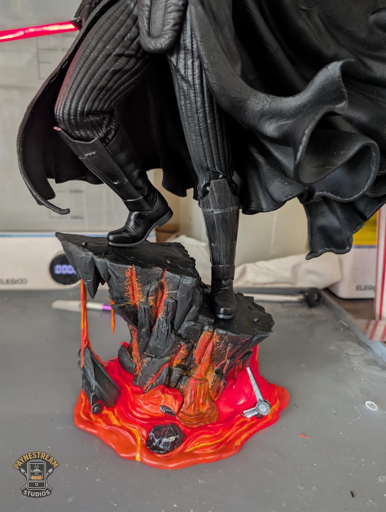 May include: A detailed figurine of a character wearing black ribbed trousers, a flowing black cape, and black boots, standing on a textured rock base with orange and red lava accents, featuring a glowing red lightsaber blade extending from the side.