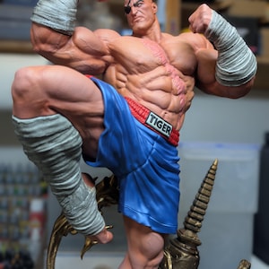 May include: A muscular, bald figurine with a black eye patch, wearing blue boxing shorts with a red waistband labeled TIGER. The figure has grey hand and leg wraps, a prominent chest scar, and is posed in a dynamic fighting stance.