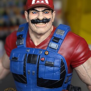 May include: A detailed figurine of a character wearing a red cap with a white "M" logo, a red shirt, and blue overalls. The character has a large black mustache and is smiling. The figurine has a realistic, muscular build and is a collectible item.