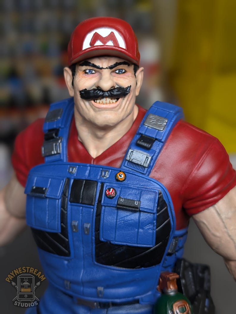 May include: A detailed figurine of a character wearing a red cap with a white "M" logo, a red shirt, and blue overalls. The character has a large black mustache and is smiling. The figurine has a realistic, muscular build and is a collectible item.