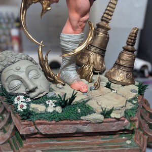 May include: A detailed figurine base featuring a stone-colored Buddha head, two weathered gold-toned temple spires, and a central figure with a wrapped foot. The base is decorated with green moss, small white flowers, and stone tiles. A logo reads Paynestream Studios.