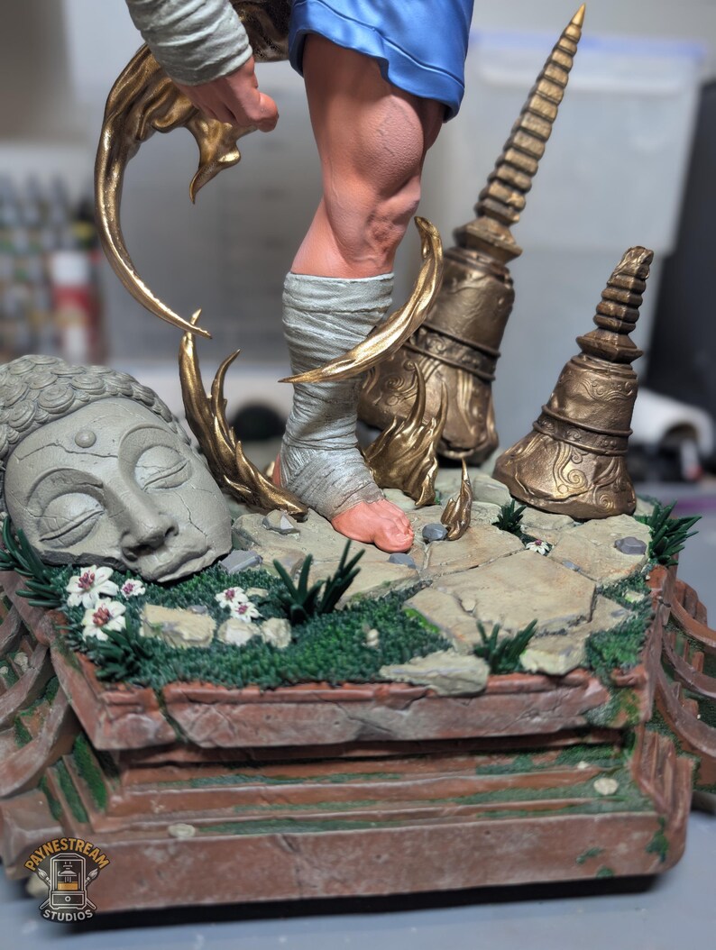 May include: A detailed figurine base featuring a stone-colored Buddha head, two weathered gold-toned temple spires, and a central figure with a wrapped foot. The base is decorated with green moss, small white flowers, and stone tiles. A logo reads Paynestream Studios.