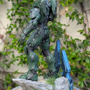May include: A Master Chief action figure standing on a rocky base with green grass and blue structural elements. The figure wears green armor with a gold visor and a backpack. A logo for Paynestream Studios is visible in the bottom left corner.