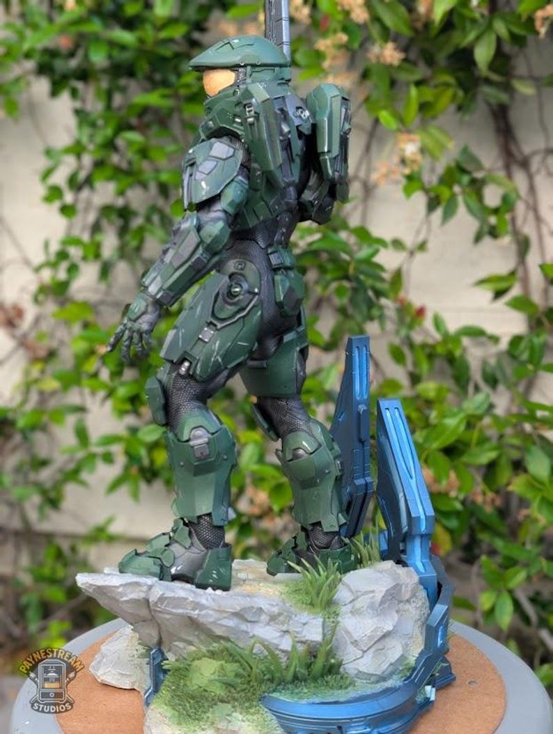 May include: A Master Chief action figure standing on a rocky base with green grass and blue structural elements. The figure wears green armor with a gold visor and a backpack. A logo for Paynestream Studios is visible in the bottom left corner.