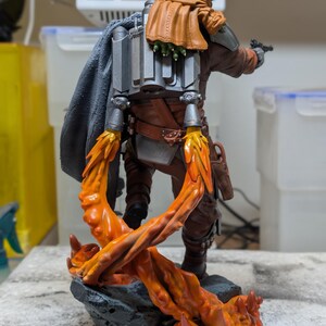 K&ouml;nnte beinhalten: Detaillierte Figur einer Figur aus einer Science-Fiction-Serie, mit silbernem Helm, brauner und grauer R&uuml;stung und einem Jetpack. Die Figur posiert mit einem Blaster, und die Basis hat orangefarbene Flammeneffekte.