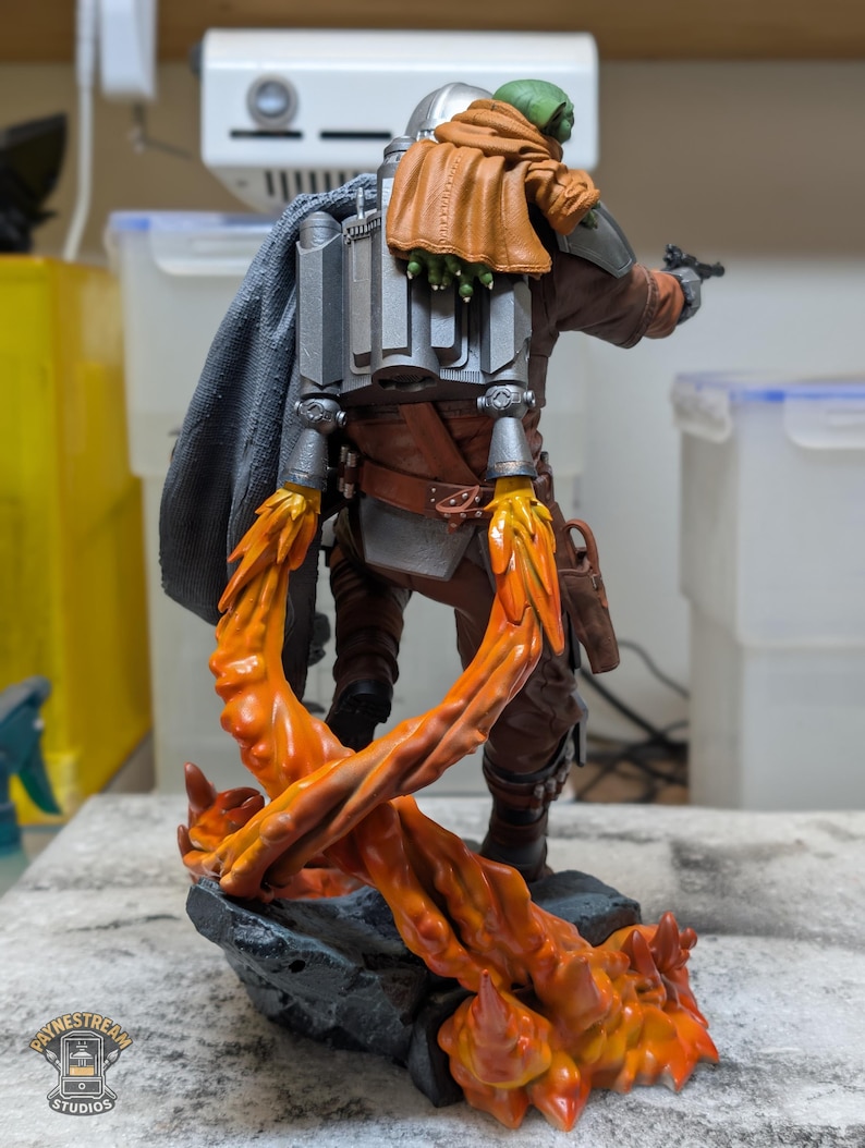 K&ouml;nnte beinhalten: Detaillierte Figur einer Figur aus einer Science-Fiction-Serie, mit silbernem Helm, brauner und grauer R&uuml;stung und einem Jetpack. Die Figur posiert mit einem Blaster, und die Basis hat orangefarbene Flammeneffekte.