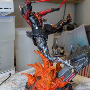 K&ouml;nnte beinhalten: Eine detaillierte Actionfigur eines geh&ouml;rnten Charakters mit roter und schwarzer Haut, bekleidet mit schwarzer Kleidung, der zwei rote Lichtschwerter f&uuml;hrt. Die Figur ist in einem dynamischen Sprung &uuml;ber einer strukturierten Basis aus orangefarbenem und schwarzem Lavagestein dargestellt.