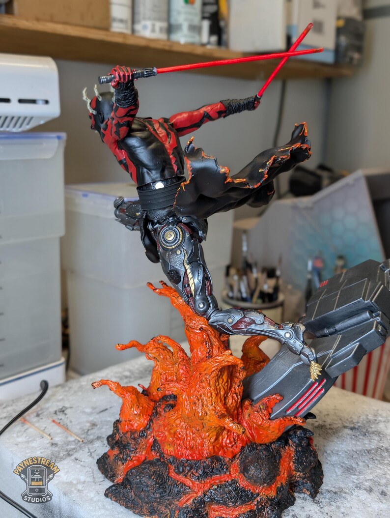K&ouml;nnte beinhalten: Eine detaillierte Actionfigur eines geh&ouml;rnten Charakters mit roter und schwarzer Haut, bekleidet mit schwarzer Kleidung, der zwei rote Lichtschwerter f&uuml;hrt. Die Figur ist in einem dynamischen Sprung &uuml;ber einer strukturierten Basis aus orangefarbenem und schwarzem Lavagestein dargestellt.