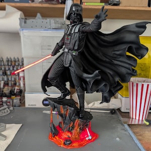 May include: A Darth Vader figurine standing on a rocky base with flowing orange and red lava. The character wears a black suit, helmet, and cape, with one hand extended forward and the other holding a glowing red lightsaber blade.