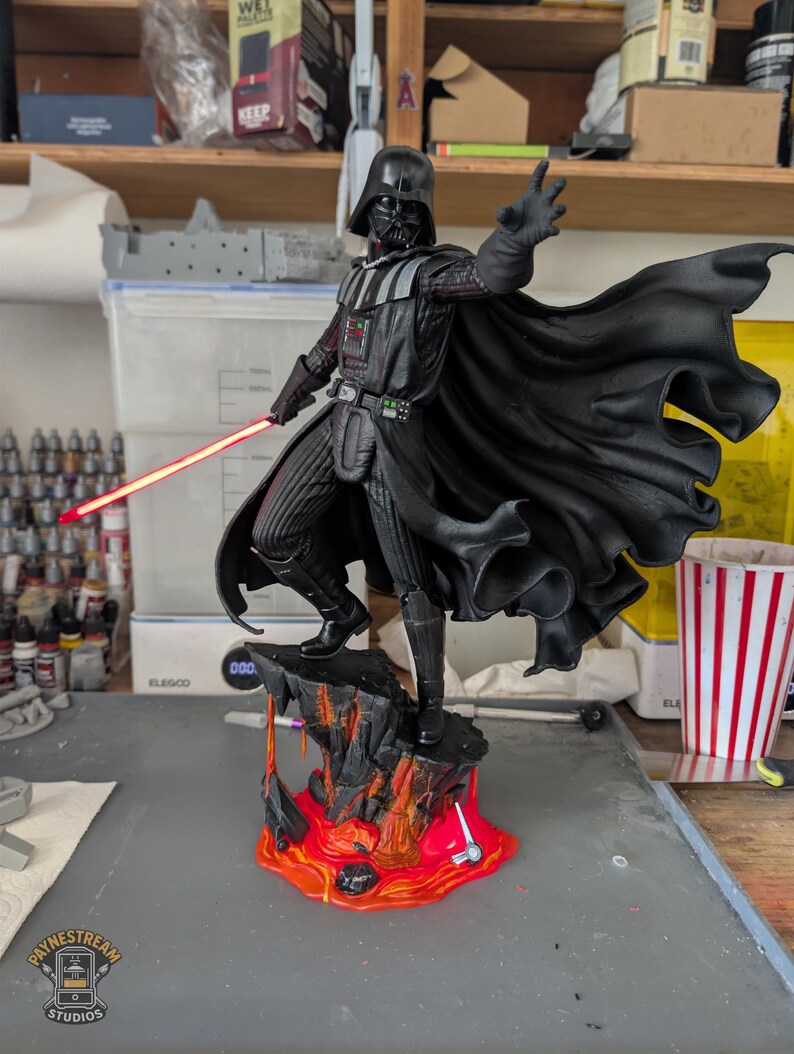 May include: A Darth Vader figurine standing on a rocky base with flowing orange and red lava. The character wears a black suit, helmet, and cape, with one hand extended forward and the other holding a glowing red lightsaber blade.