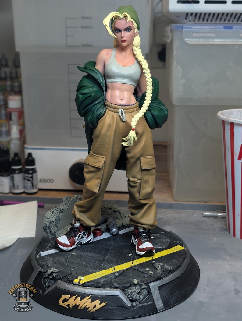 May include: A collectible figurine of a blonde character with a long braid, wearing a green beanie, a light gray crop top, a dark green jacket, and tan cargo pants. The figure stands on a circular base with a street design and the text CAMMY.