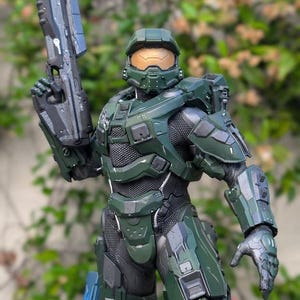 May include: A detailed action figure of a soldier in green armor with a gold visor, holding a large dark gray rifle. The armor features a textured black undersuit, segmented plating, and a logo for Paynestream Studios on the chest piece.