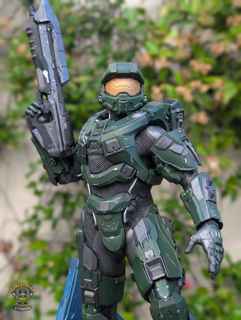 May include: A detailed action figure of a soldier in green armor with a gold visor, holding a large dark gray rifle. The armor features a textured black undersuit, segmented plating, and a logo for Paynestream Studios on the chest piece.