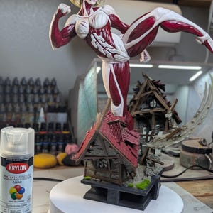 May include: A hand-painted figurine of a blonde female titan with exposed red muscle tissue and white bone-like armor, captured in a high-kick pose atop a ruined, shingled house structure. A can of Krylon ColorMaxx clear spray paint stands nearby.